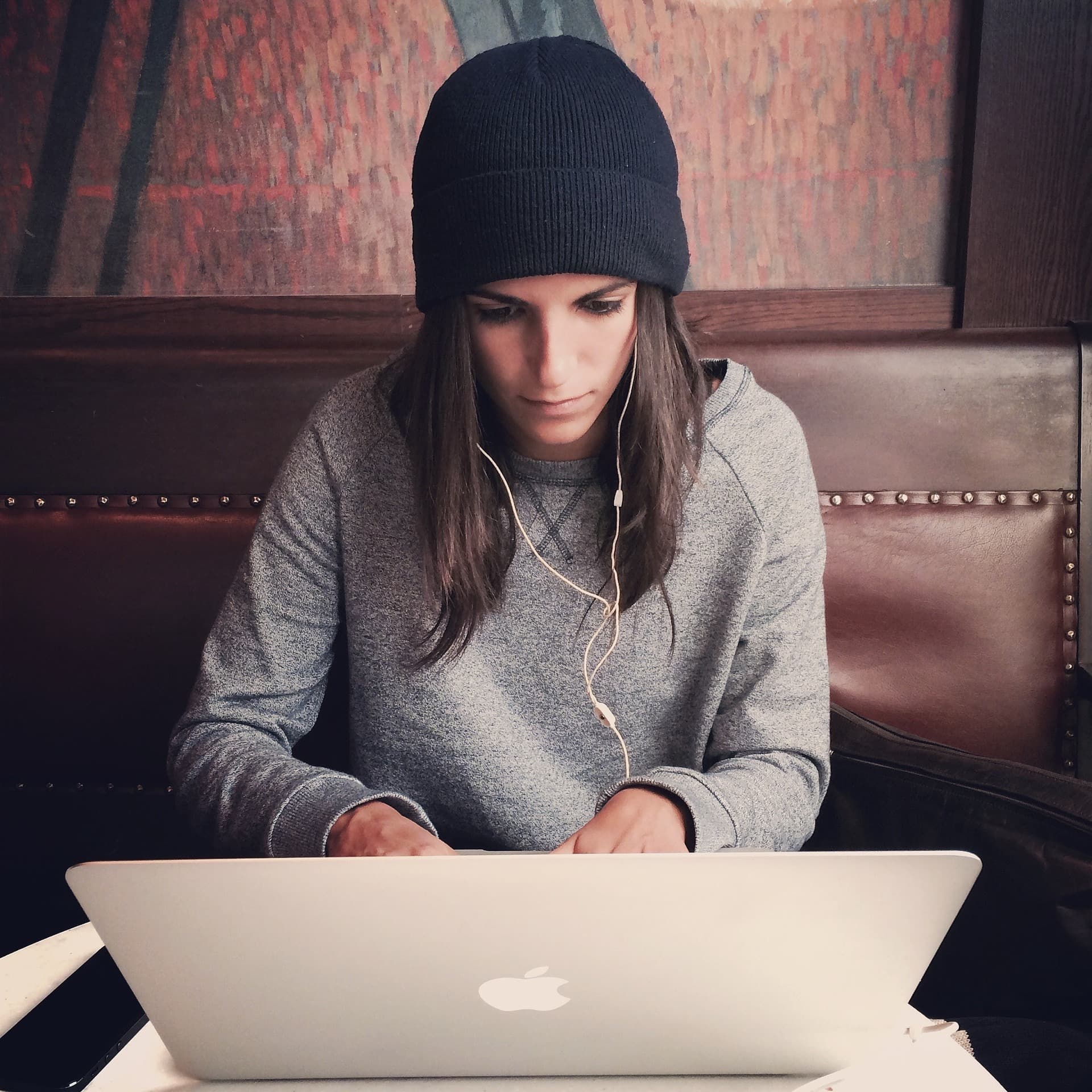 A young woman with dark brown long hair wearing a navy blue beanie hat sitting working at a laptop.